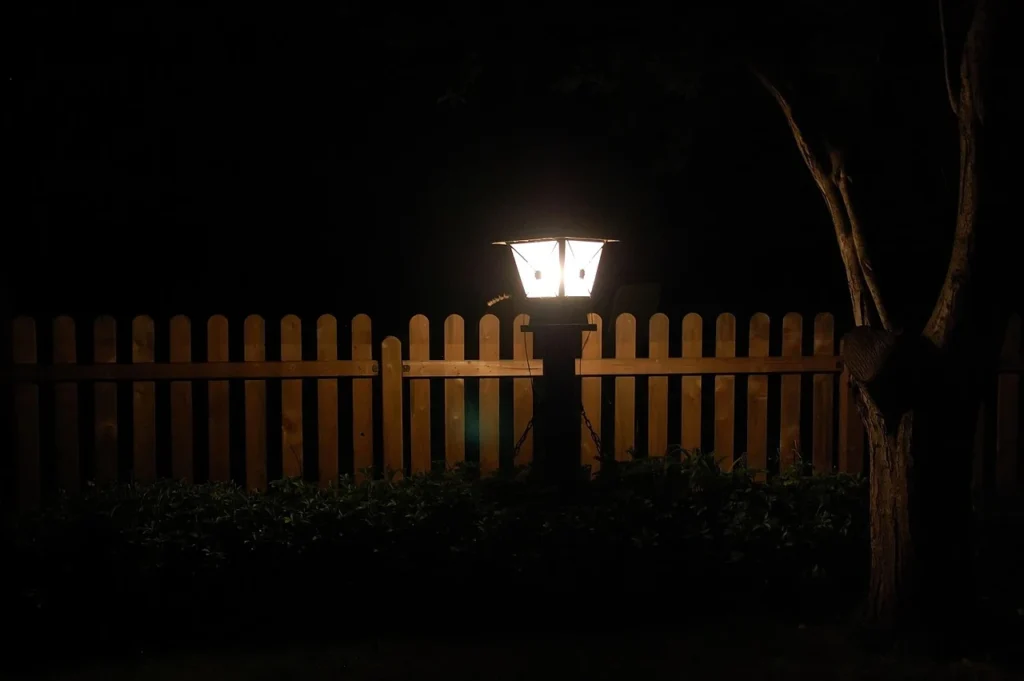 Lampa ogrodowa przy drewnianym płocie nocą – punktowe oświetlenie ścieżki w ogrodzie.