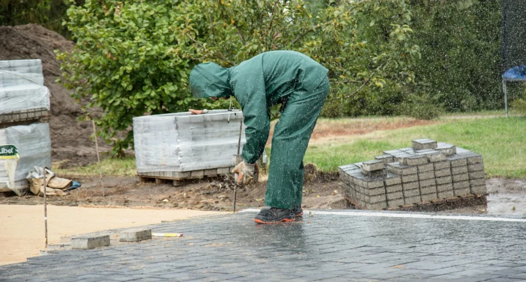 Brukarz w zielonym komplecie przeciwdeszczowym układa kostkę brukową podczas deszczu.