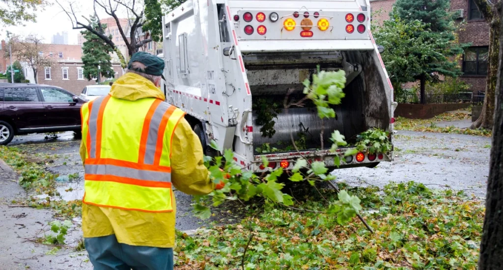 Pracownik służb komunalnych w kamizelce odblaskowej sprząta liście przy śmieciarce.