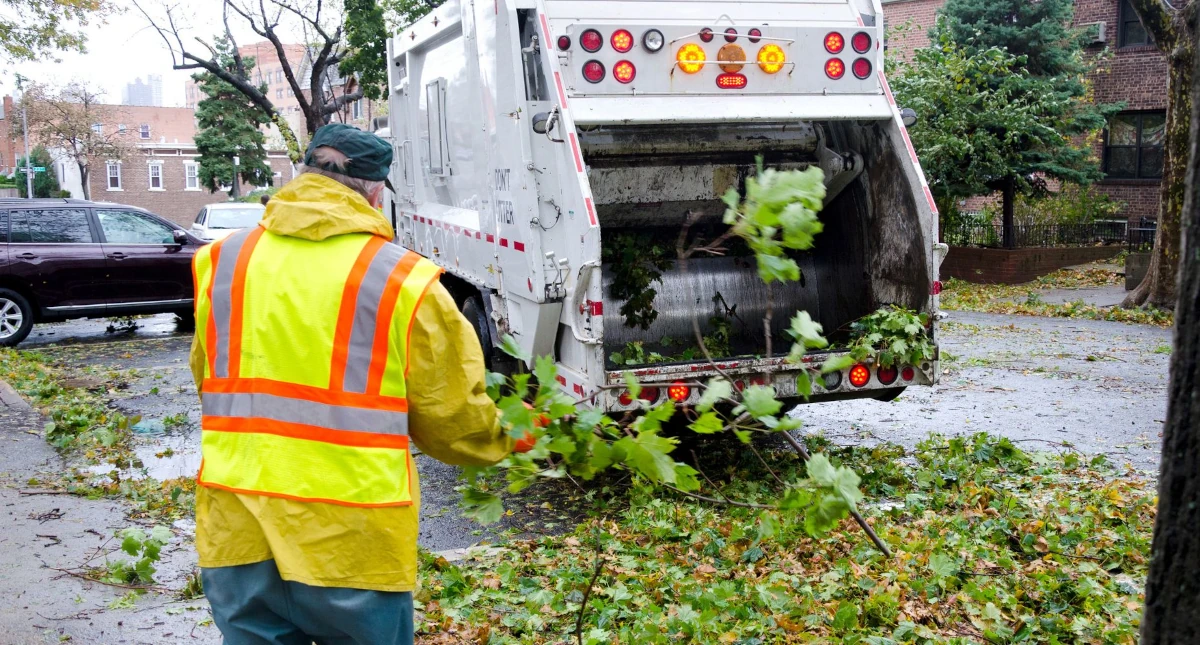 Pracownik służb komunalnych w kamizelce odblaskowej sprząta liście przy śmieciarce.